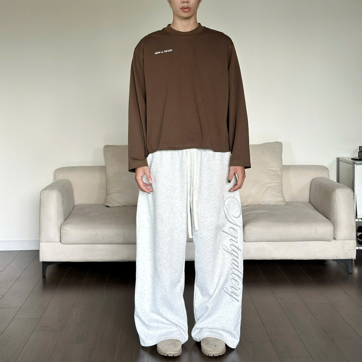Person wearing a brown long-sleeve shirt and white pants standing in a living room.