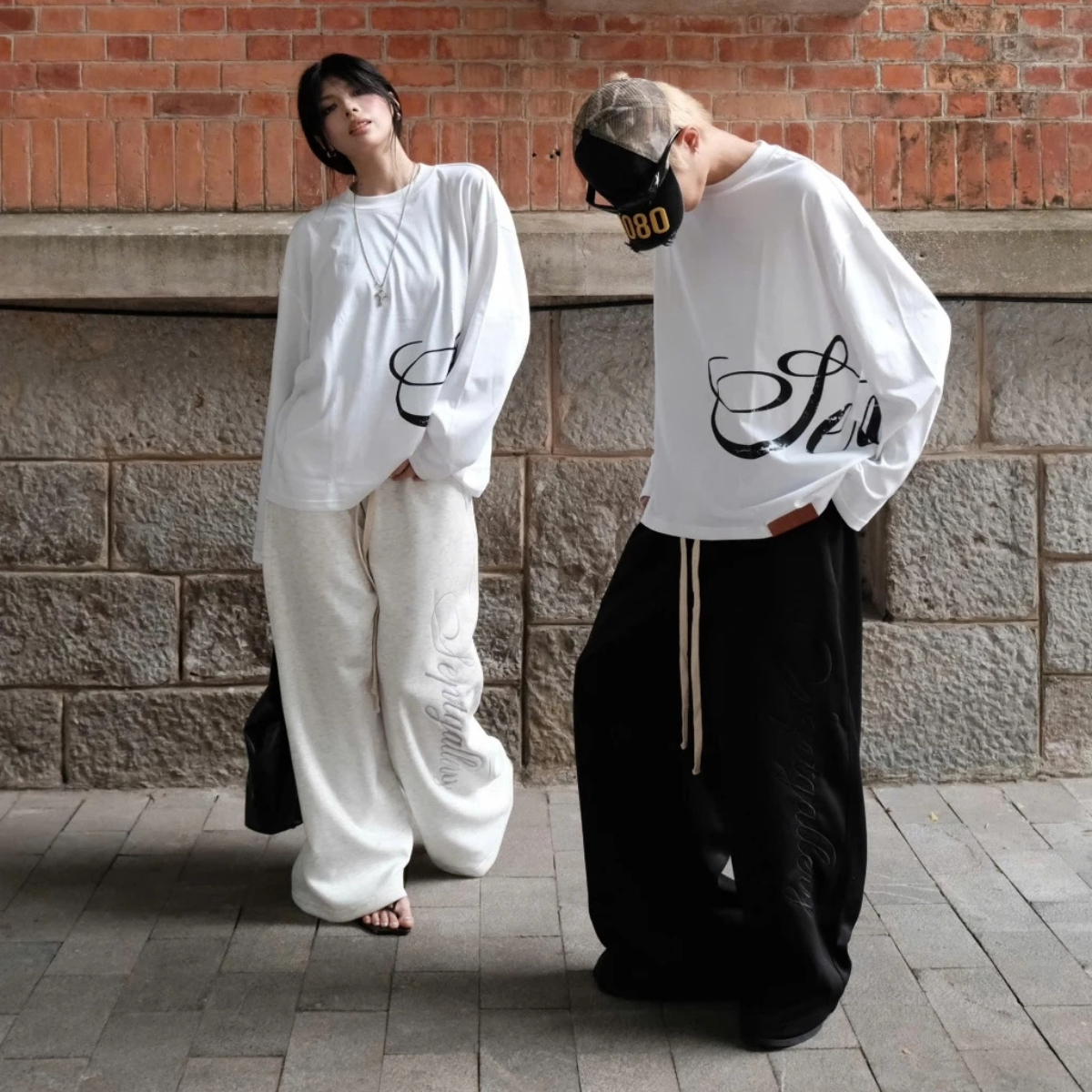 Two people wearing white and black outfits with text designs, standing against a brick wall.