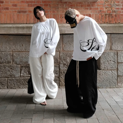 Two people wearing white and black outfits with text designs, standing against a brick wall.