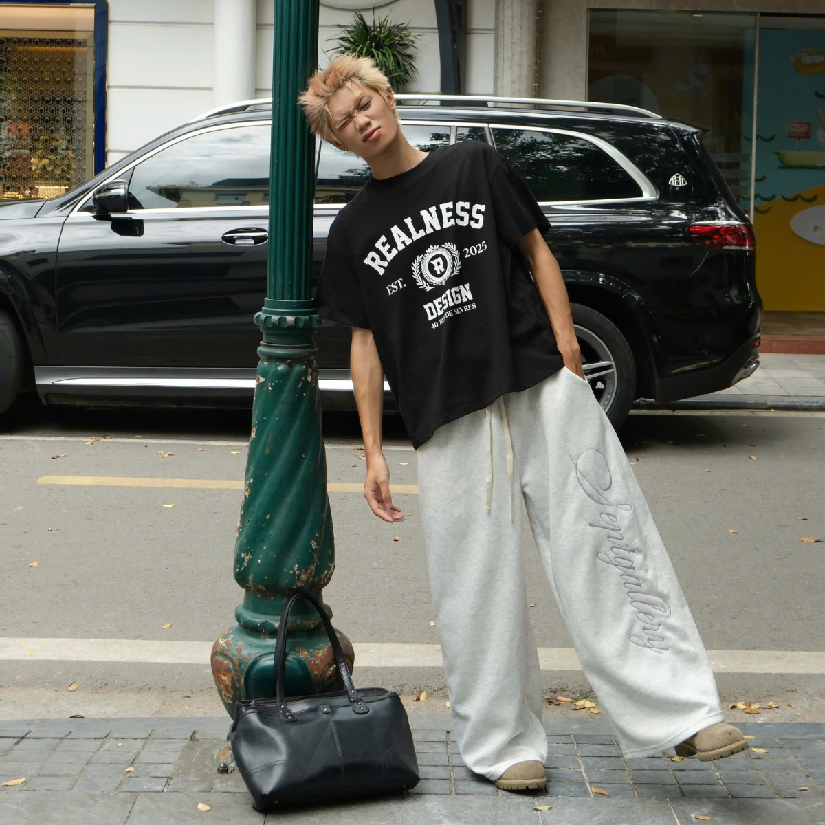 Person wearing a black 'REALNESS' t-shirt and light gray pants standing on a street next to a black SUV.