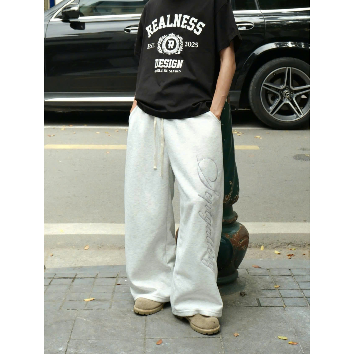 Person wearing a black 'Realness' t-shirt and white pants standing on a street.