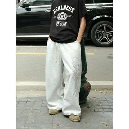 Person wearing a black 'Realness' t-shirt and white pants standing on a street.