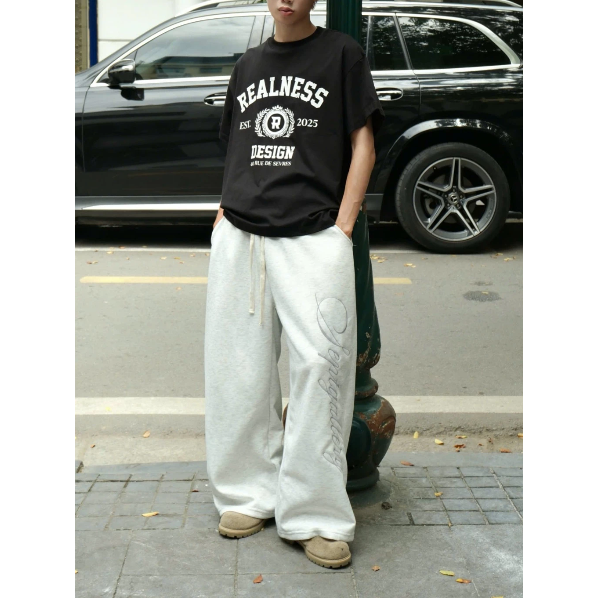 Person wearing a black 'REALNESS' t-shirt and white pants standing on a street.