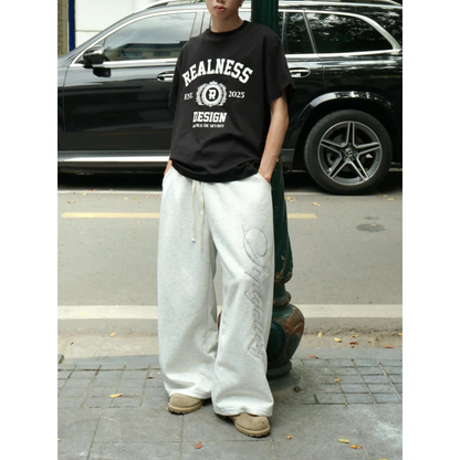 Person wearing a black 'REALNESS' t-shirt and white pants standing on a street.